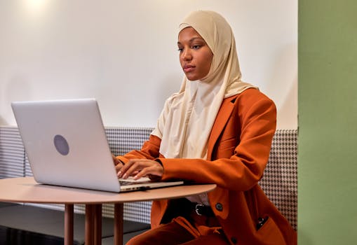 Confident woman in a hijab using a laptop in a modern office setting.